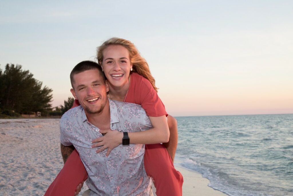 young man giving fiancée a piggy back ride on the beach at sunset