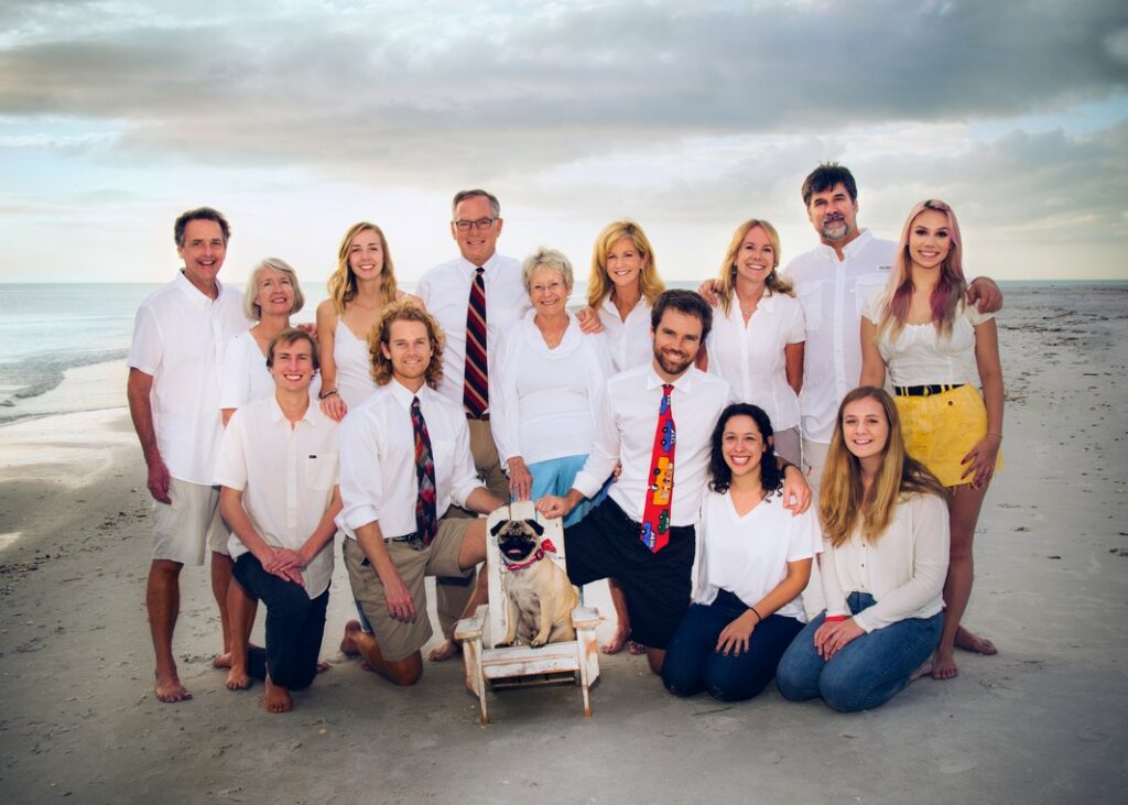 large family grouped around a pug dog sitting in a white adirondack chair on the beach